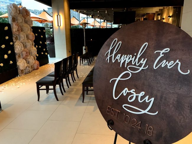 picture of Truluck's Woodlands wedding event on the Terrace with "Happily Ever After Cosey" sign at the entry and a floor to ceiling paper flower installed background for the couple to marry in front of. Chairs are setup in rows for the ceremony overlooking the water.