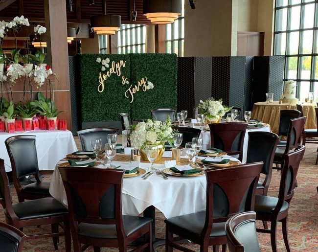 picture of Truluck's Woodlands private dining event featuring orchid take away gifts, a beautiful green hedge with the bride and grooms names made out of wood and painted in gold to hang on the hedge. In the corner is a cake table dressed in gold sequin linen with a 3 tiered white wedding cake. Table accents are gold with deep forest green and white roses.