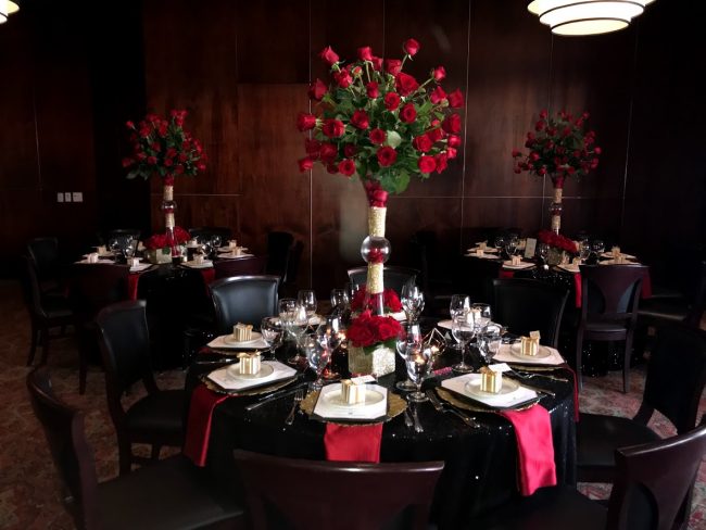 picture of Truluck's Woodlands private event with very dramatic black sequin linens with red satin accents including tall floral centerpieces full of red roses with a take away box of chocolates.