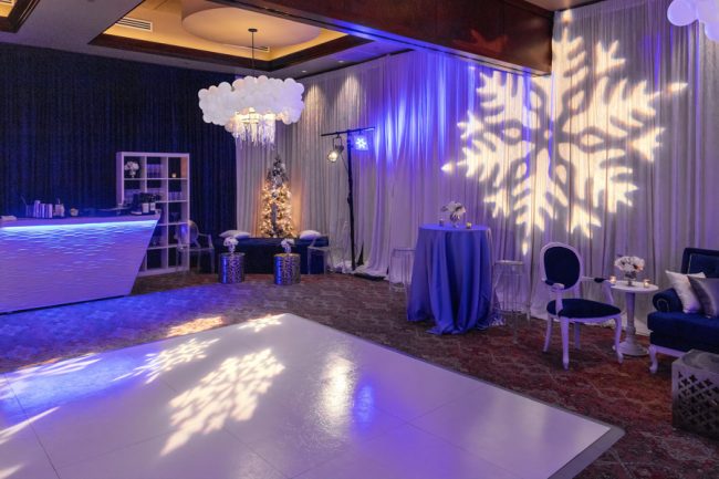picture of Truluck's Woodlands Winter Wonderland holiday celebration to include a white dance floor, white bar that is lit in bluish light. The ceiling lamps were dressed in silver tinsel and white balloons. Accent furniture in royal blue and white was brought in and snowflake gobos covered the space. The walls were draped in royal blue and white with up lighting and a silver and white Christmas tree is in the background.