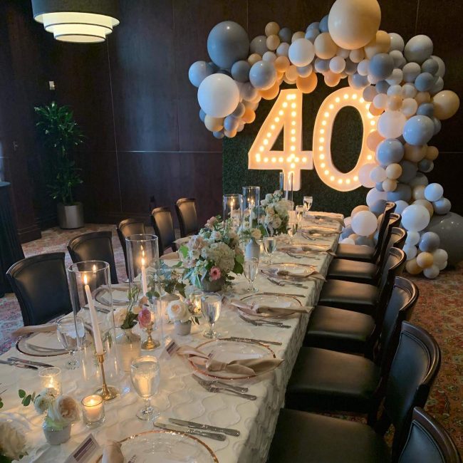 picture of Truluck's Woodlands private dining event for a 40th birthday celebration. This includes white and cream linens with gold accents to include gold rimmed table chargers, gold candle sticks encased in glass with white taper candles. On the back wall is a green hedge with 5' lit up "40" encased in white, gold and gray balloons.