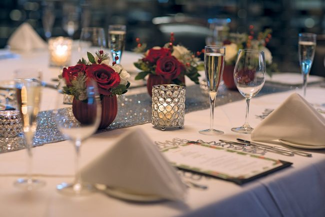picture of private dining holiday decor with silver table runner, fresh flowers that include red and white roses and holiday greenery, crystal candles, specialty menus