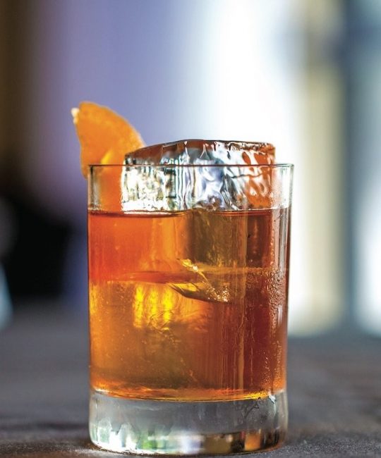 picture of Old fashioned poured in a cocktail glass with a large block of ice and an orange rind
